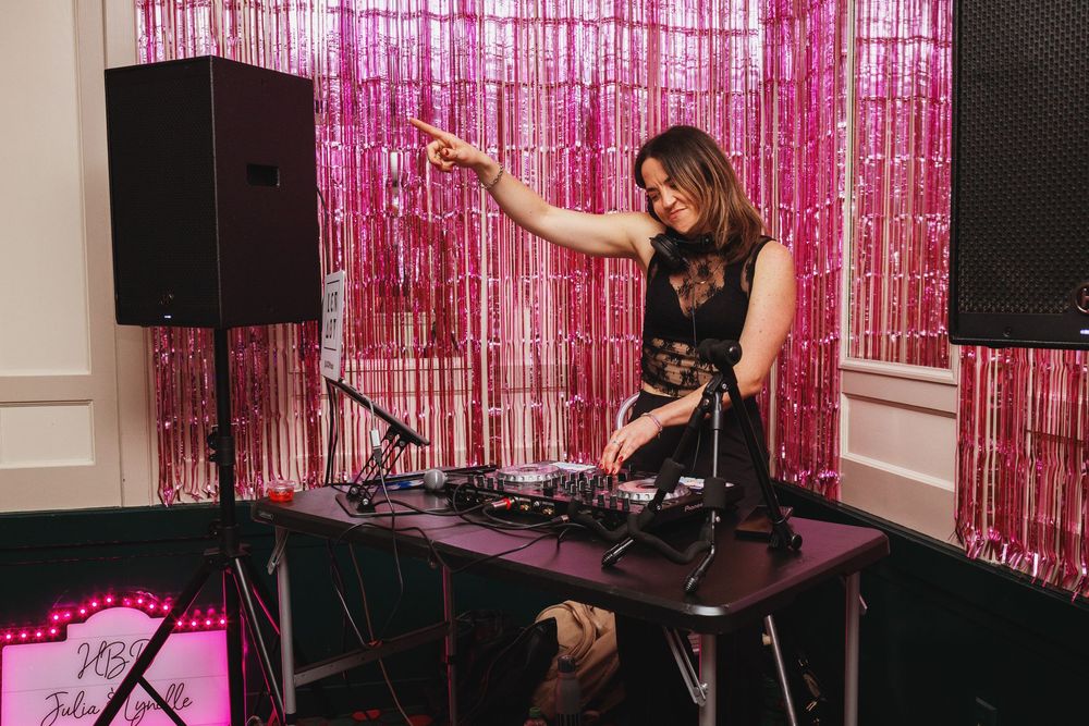 Female DJ performing at a pink tinsel backdrop party event, mixing tracks on professional equipment in elegant venue.
