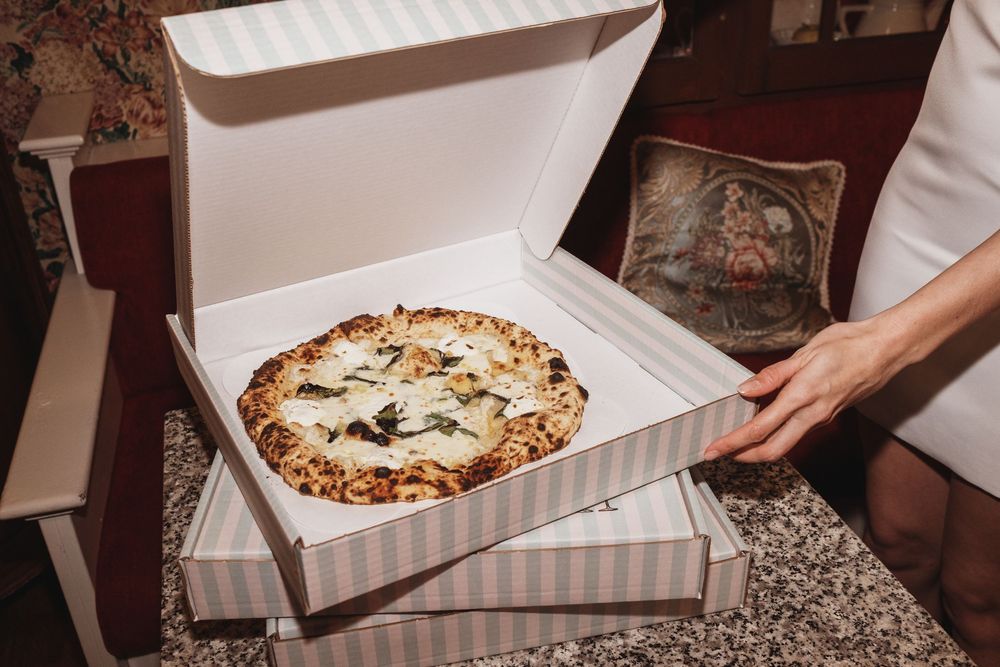 Freshly baked white pizza with mushrooms and herbs in an open striped delivery box held by hands.