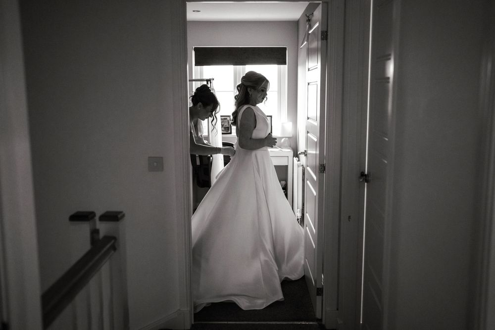 Black and white photo of a bride in a white gown being helped by a bridesmaid in a softly lit hallway.