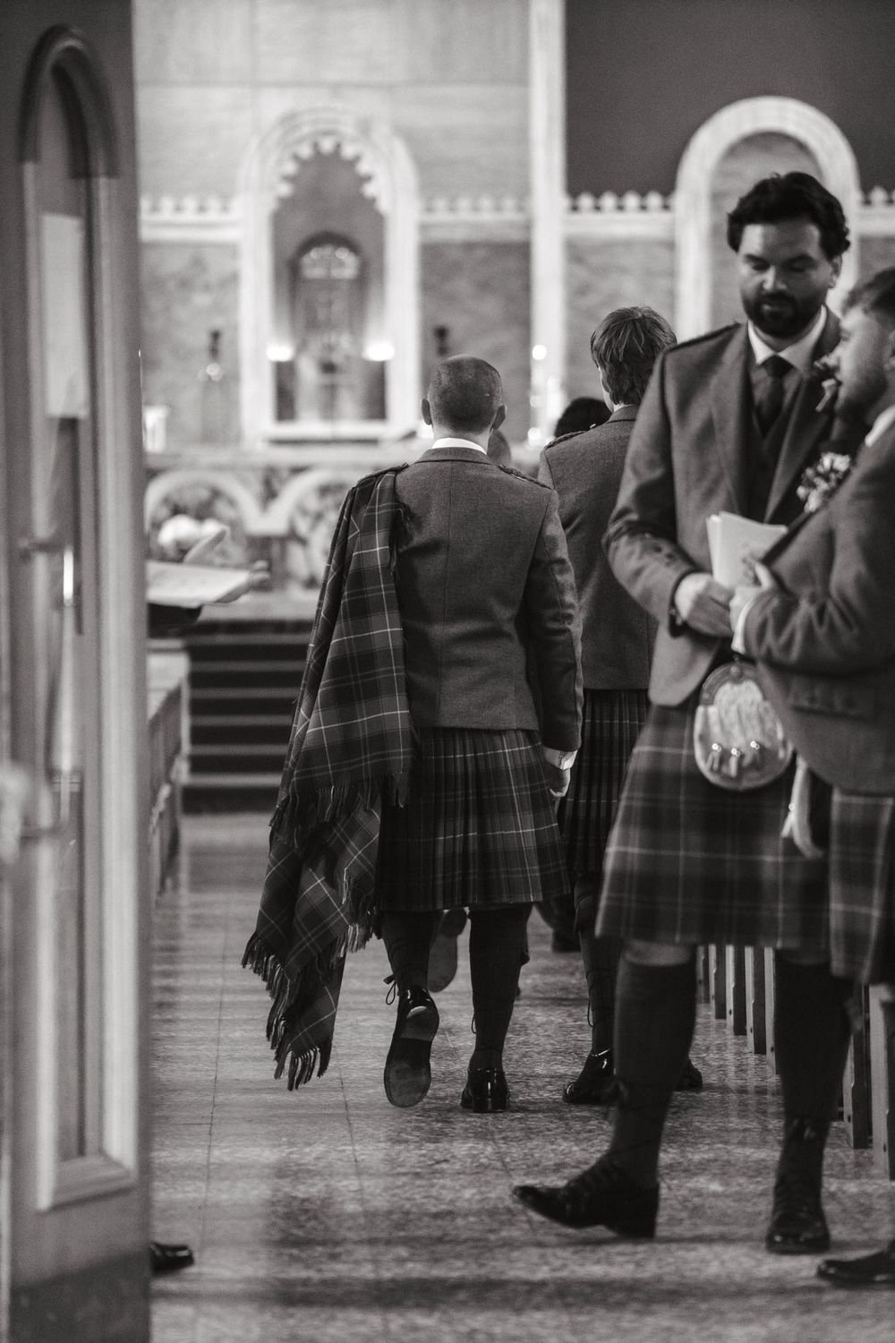 Black and white photo of men in traditional Scottish kilts walking through a church interior.
