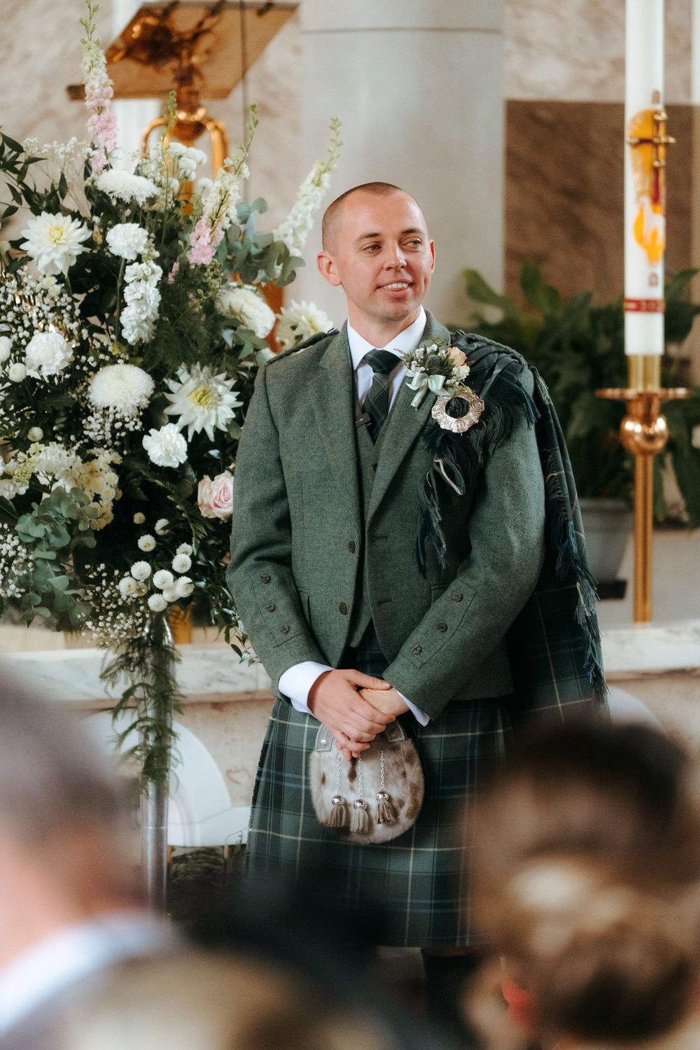 Smiling groom in green tartan kilt and jacket with boutonniere standing at church altar during wedding ceremony.