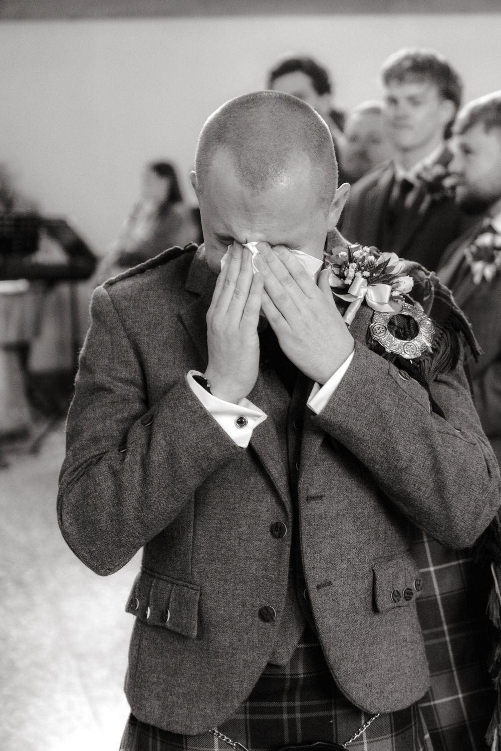 Emotional groom in tweed suit covering face during wedding ceremony, black and white photo.