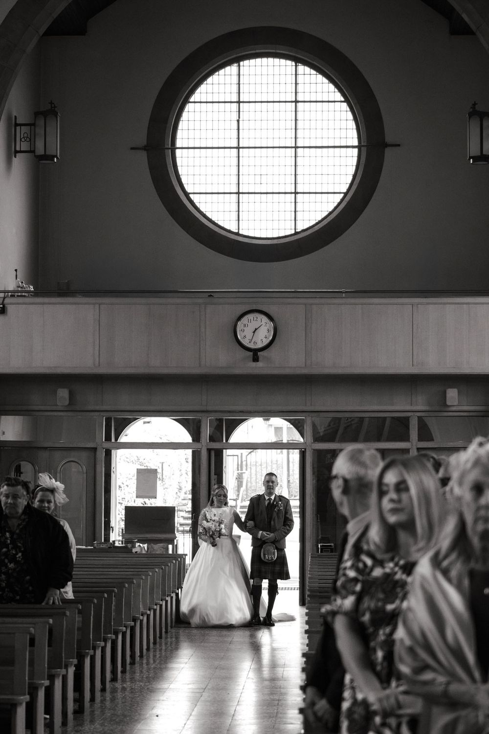 Black and white wedding photo of bride and groom walking down church aisle beneath a large circular stained glass window.