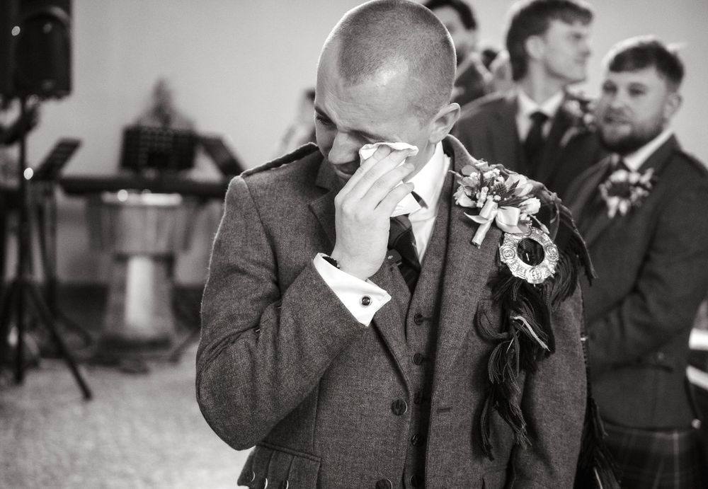 Emotional groom wiping tears at wedding ceremony in black and white photo, wearing suit with floral boutonniere.