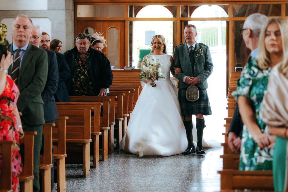 Bride in white gown walks down church aisle with man in Scottish kilt at wedding ceremony.