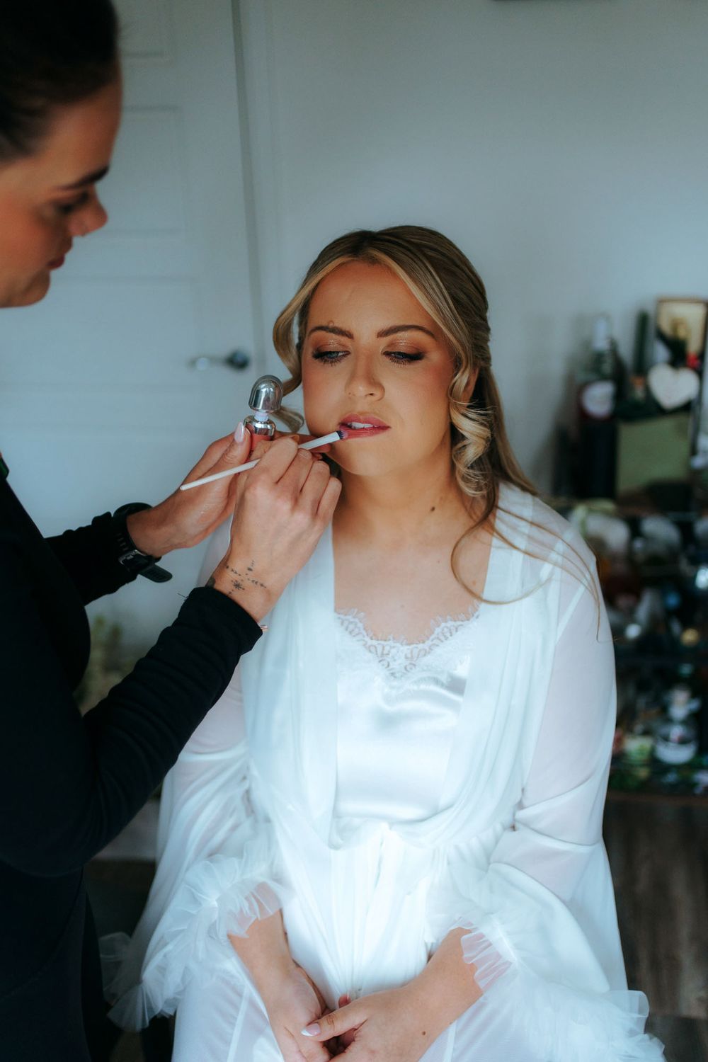 Makeup artist applying lipstick to a bride in a white robe during bridal wedding preparation.