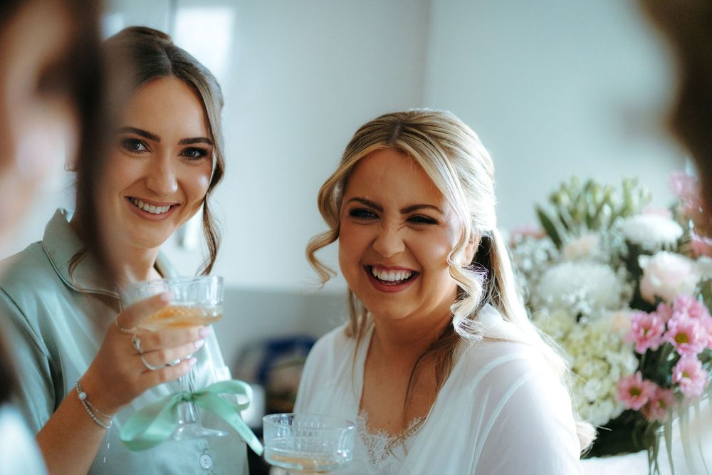 Two bridesmaids smiling and toasting with champagne glasses at a wedding celebration with floral arrangements in background.