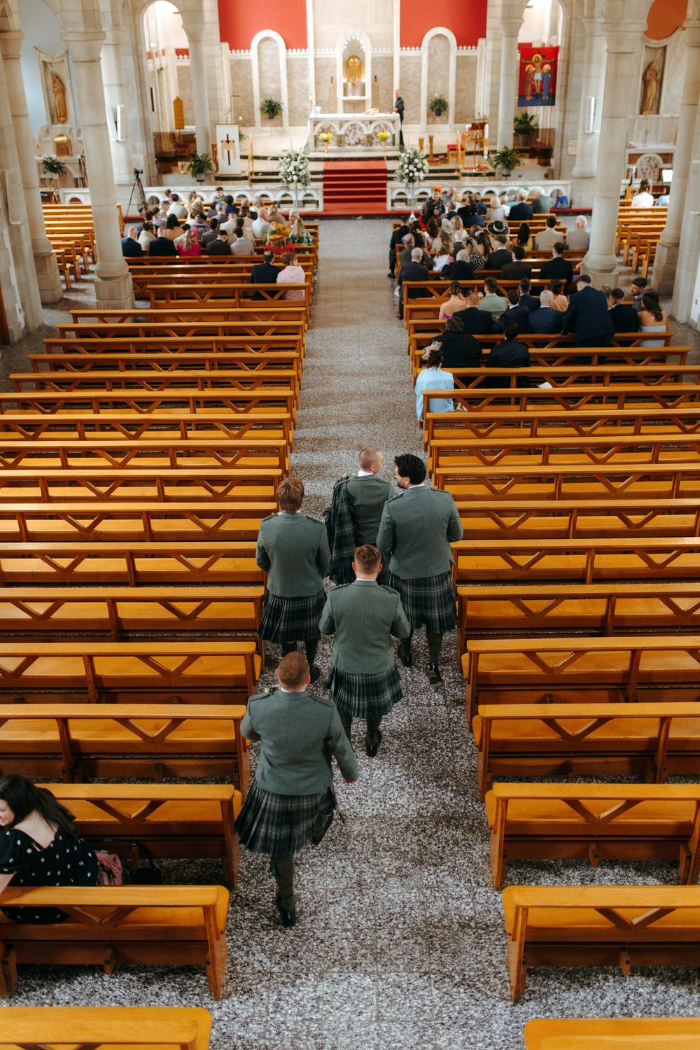 Men in kilts walking down church aisle during wedding ceremony, wooden pews filled with guests, altar in background.