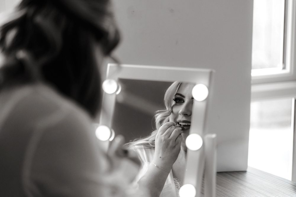 Bride applies lipstick while looking into a Hollywood mirror with lights in black and white photo.