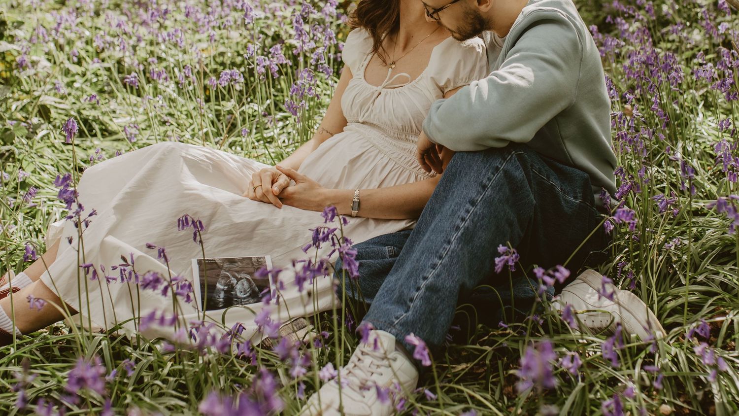 A couple sits romantically among purple wildflowers, holding hands and sharing a tender moment in a lush meadow.