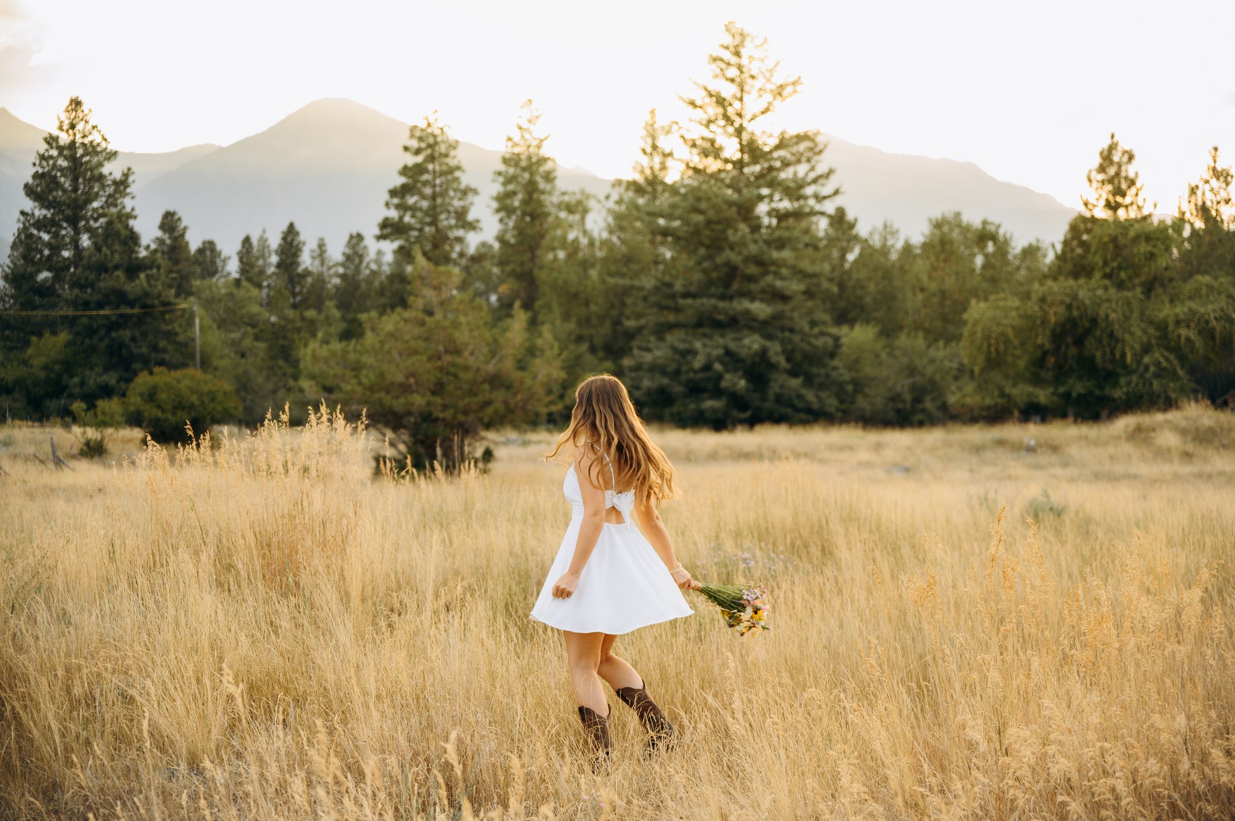Person in white dress twirls through golden grass field with mountains and evergreen trees in background at sunset.
