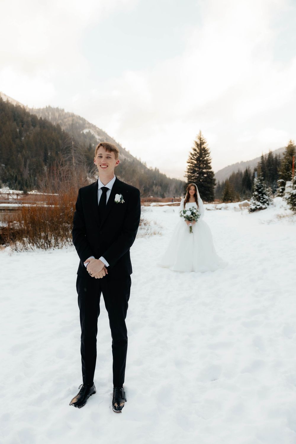 Groom in black suit smiles in snowy mountain landscape as bride in white gown holds bouquet in background.