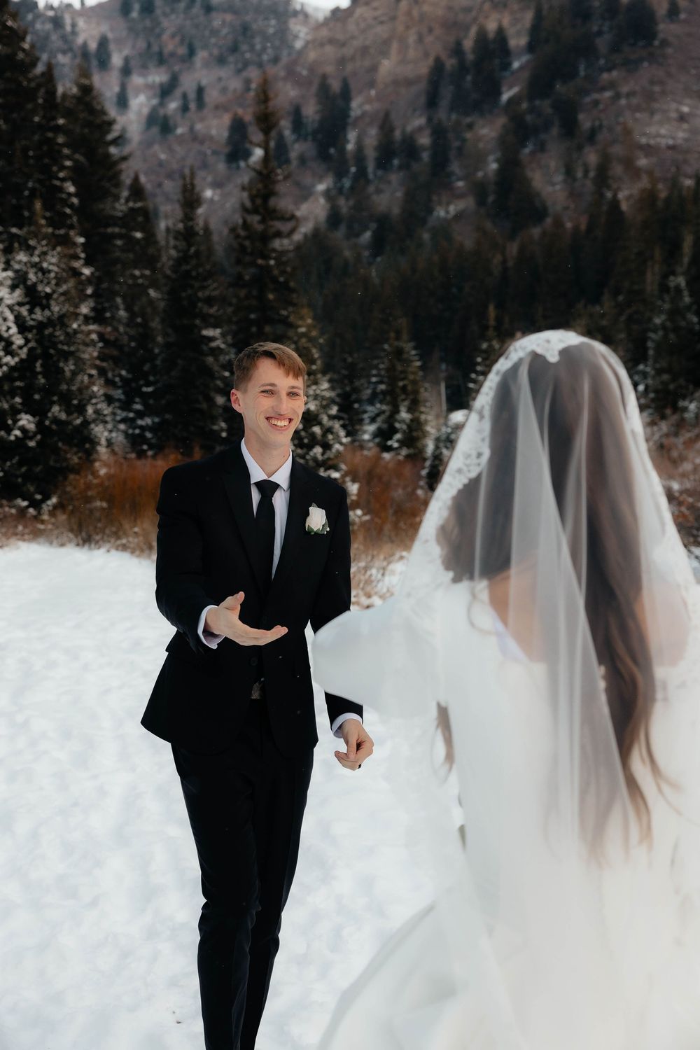 Smiling groom in black suit extends hand to bride in white gown and veil in snowy mountain forest setting.