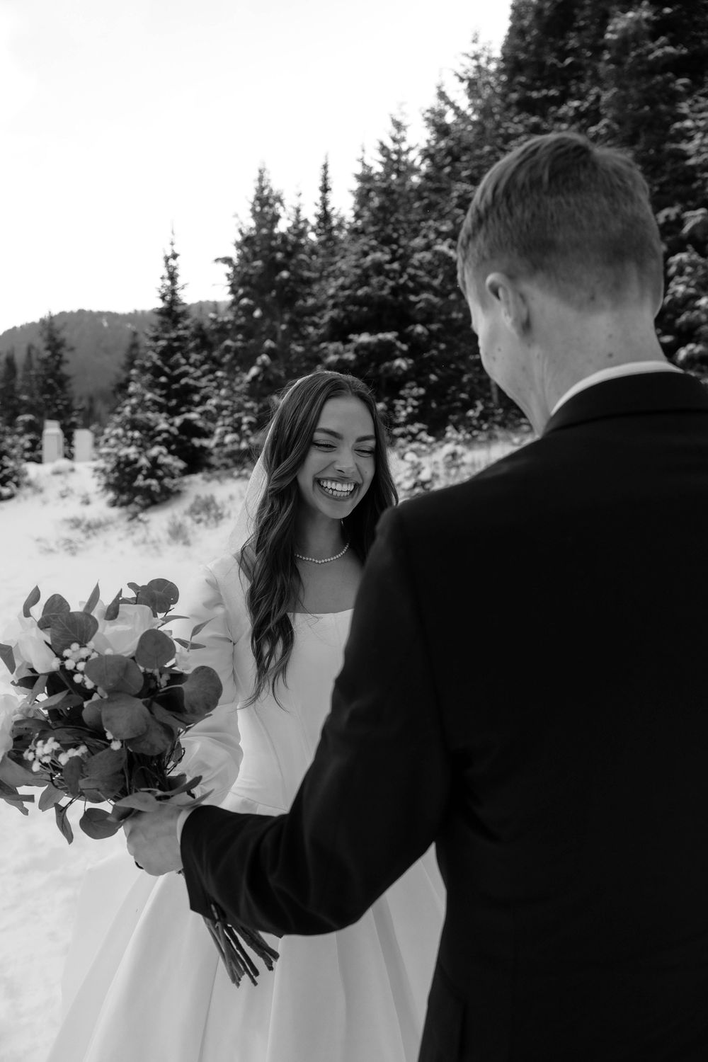 Black and white photo of radiant bride holding bouquet laughing with groom in snowy pine forest.
