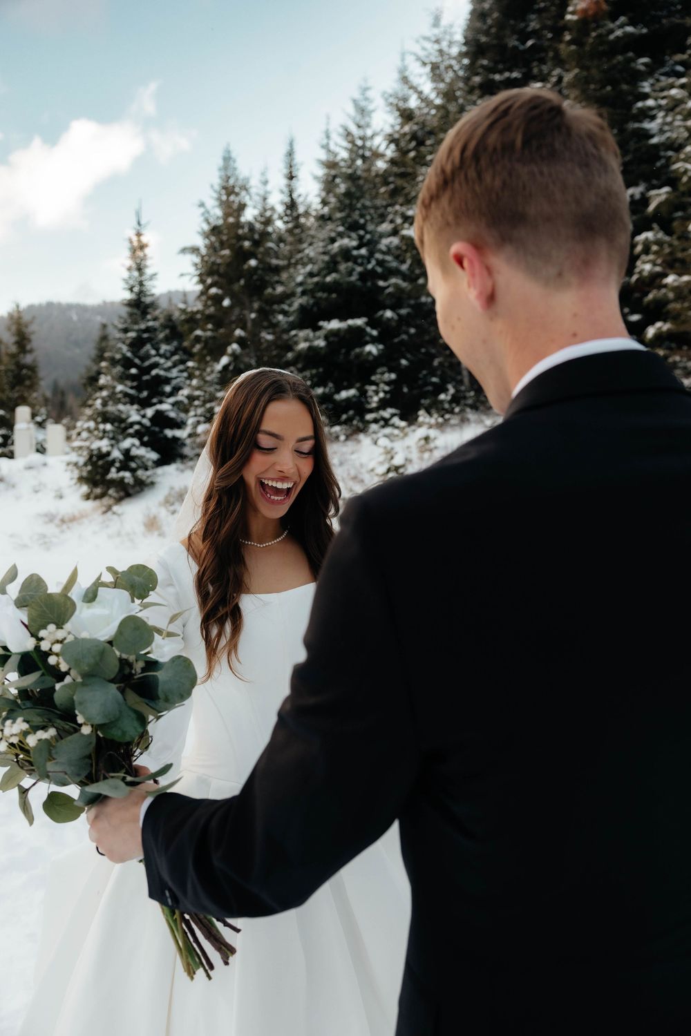 Bride in white gown laughing as groom presents eucalyptus bouquet in snowy mountain forest winter wedding.