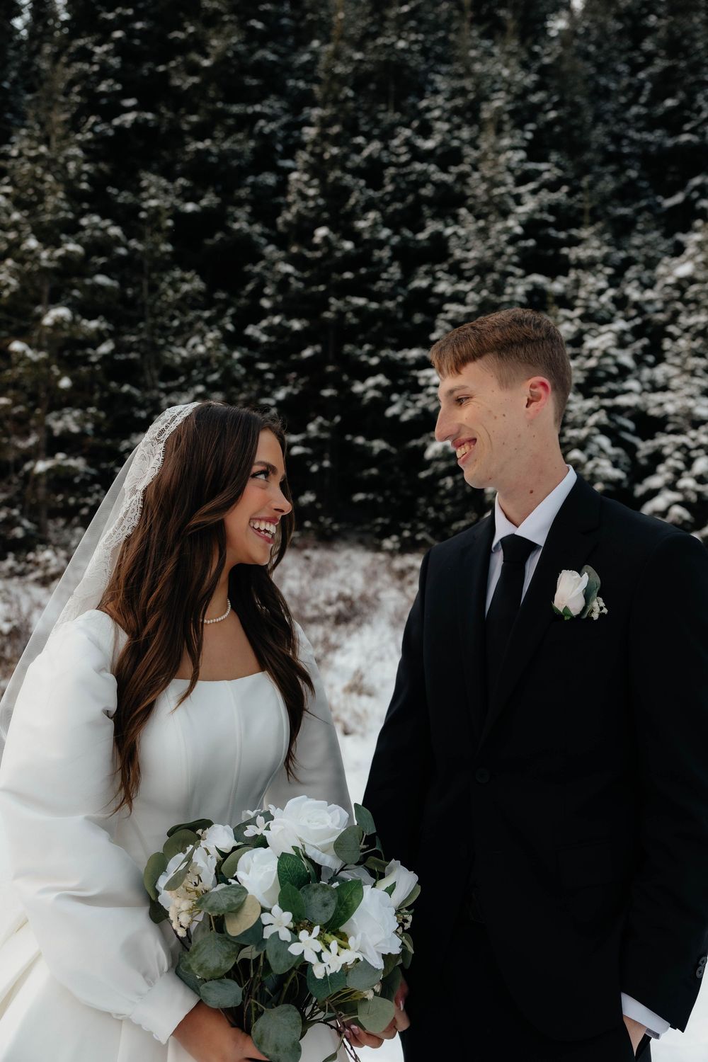 Bride and groom smiling at each other in a snowy winter forest, bride holding white rose bouquet.