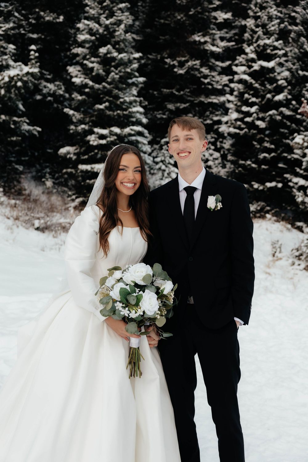 Bride and groom smile together in snowy winter forest, she holds white bouquet in elegant long-sleeve gown.