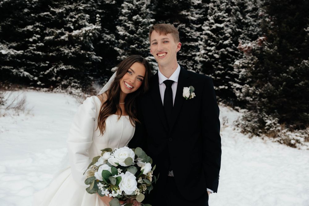 Bride and groom smiling together in snowy winter setting, bride holding white bouquet, groom in black suit with boutonniere.