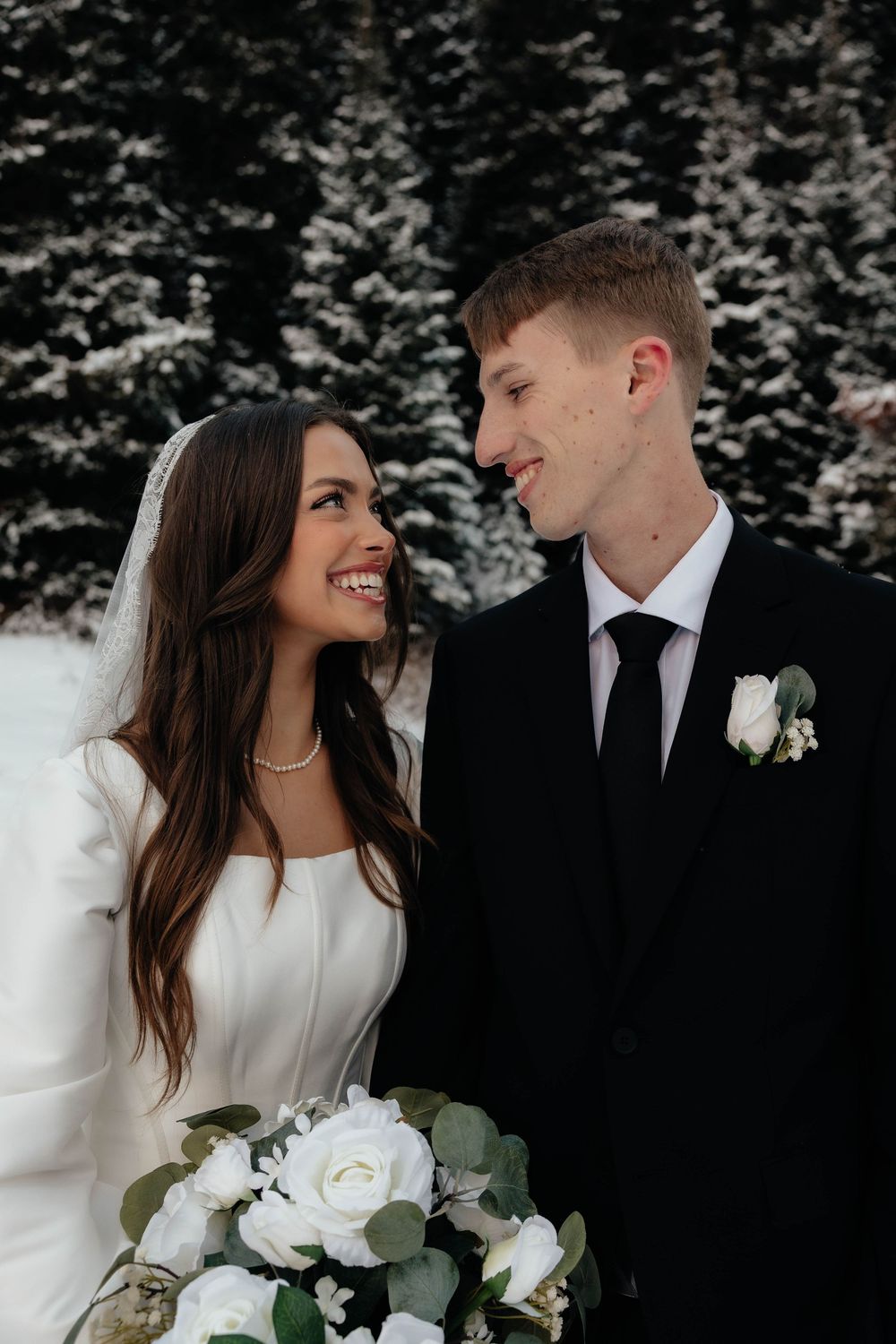 Bride and groom share a joyful moment gazing at each other in a snowy winter forest wedding setting.