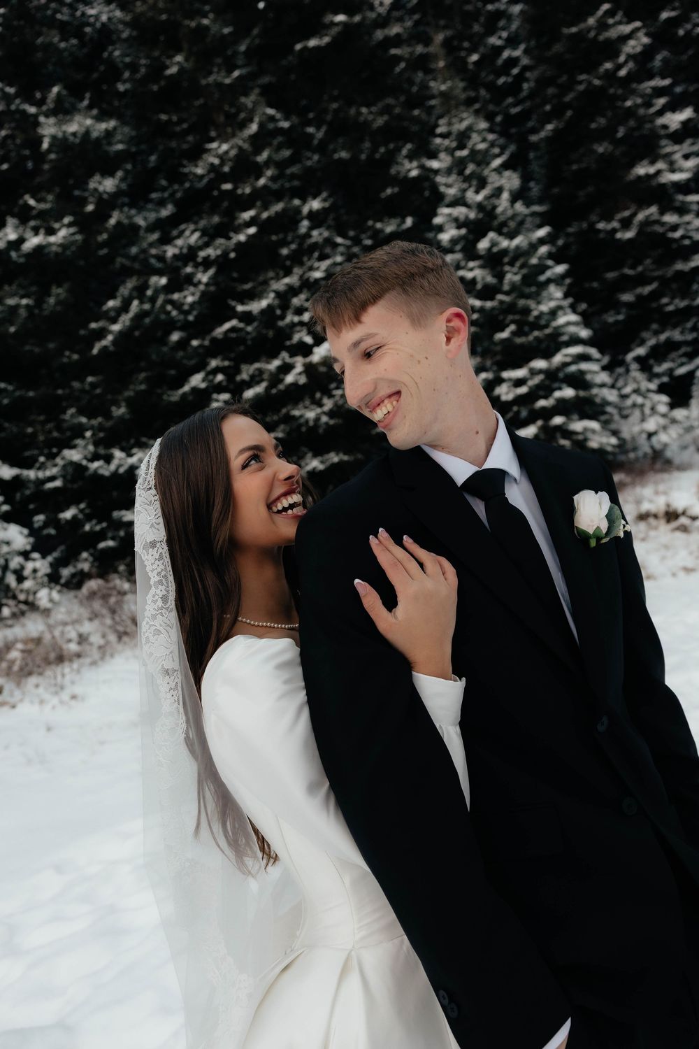 Smiling bride and groom share a joyful moment outdoors in a snowy winter wedding setting with snow-covered evergreen trees.
