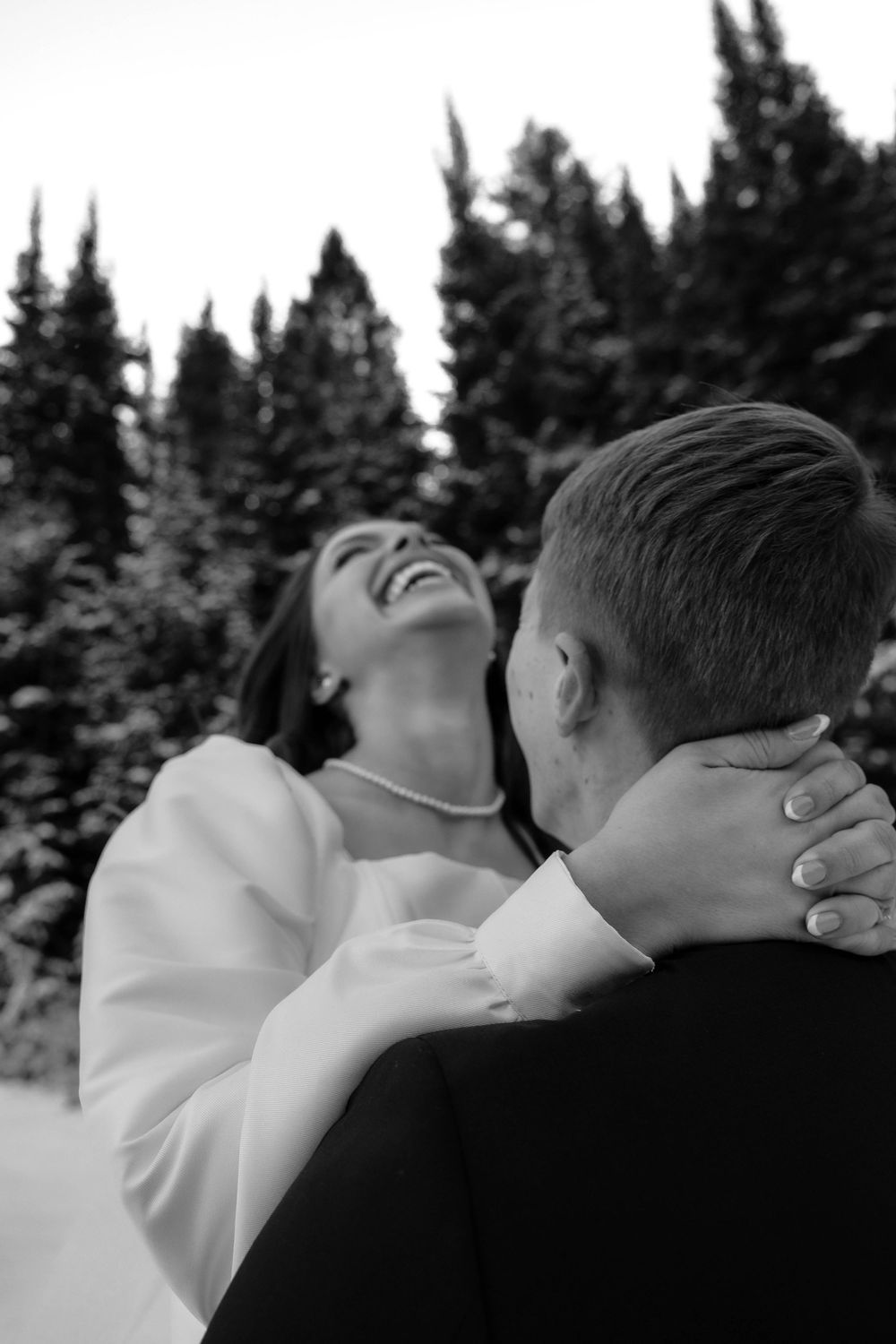 Black and white photo of laughing bride embracing groom outdoors among snow-dusted evergreen trees.
