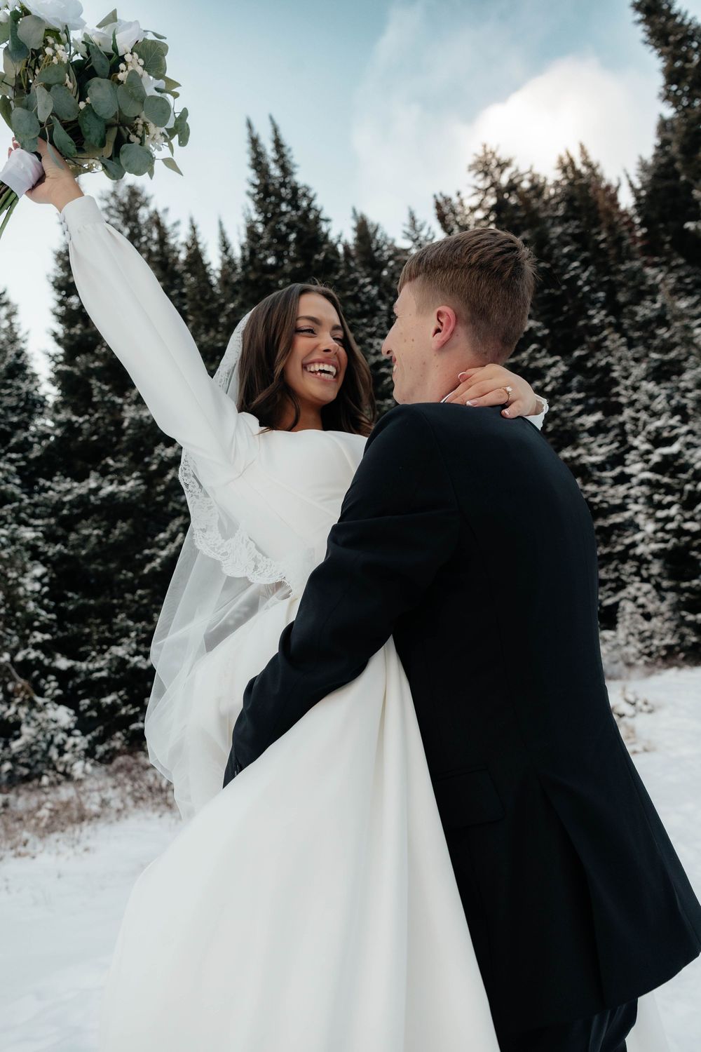 Joyful bride raises bouquet while groom lifts her in snowy winter forest wedding portrait.
