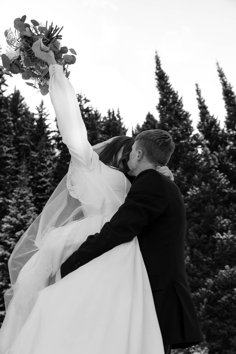 Black and white photo of a bride and groom embracing, bride raising bouquet with evergreen trees behind.