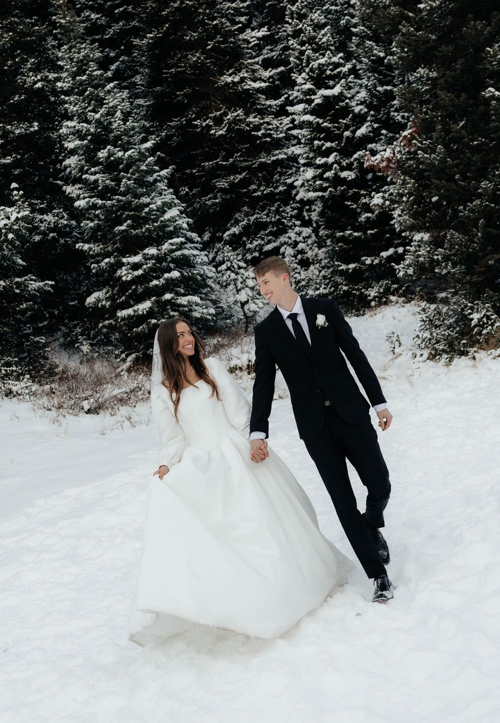 Bride and groom walking hand in hand through a snowy winter landscape with snow-covered evergreen trees.