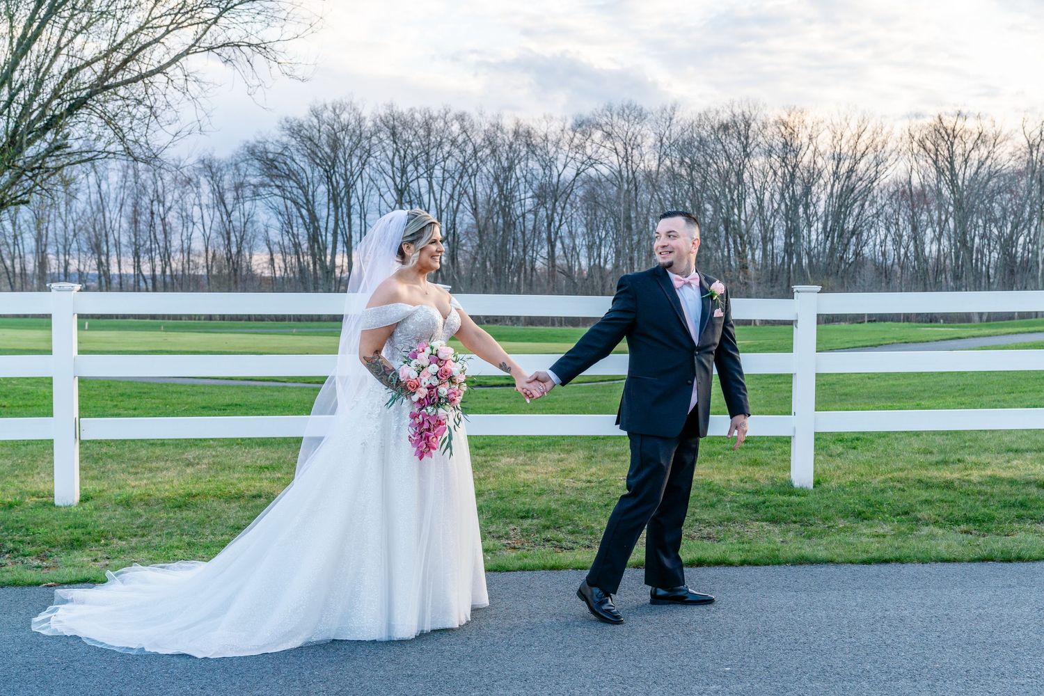 Bride and groom hold hands walking along a white fence at an outdoor wedding venue during golden hour at Black Swan Country Club.