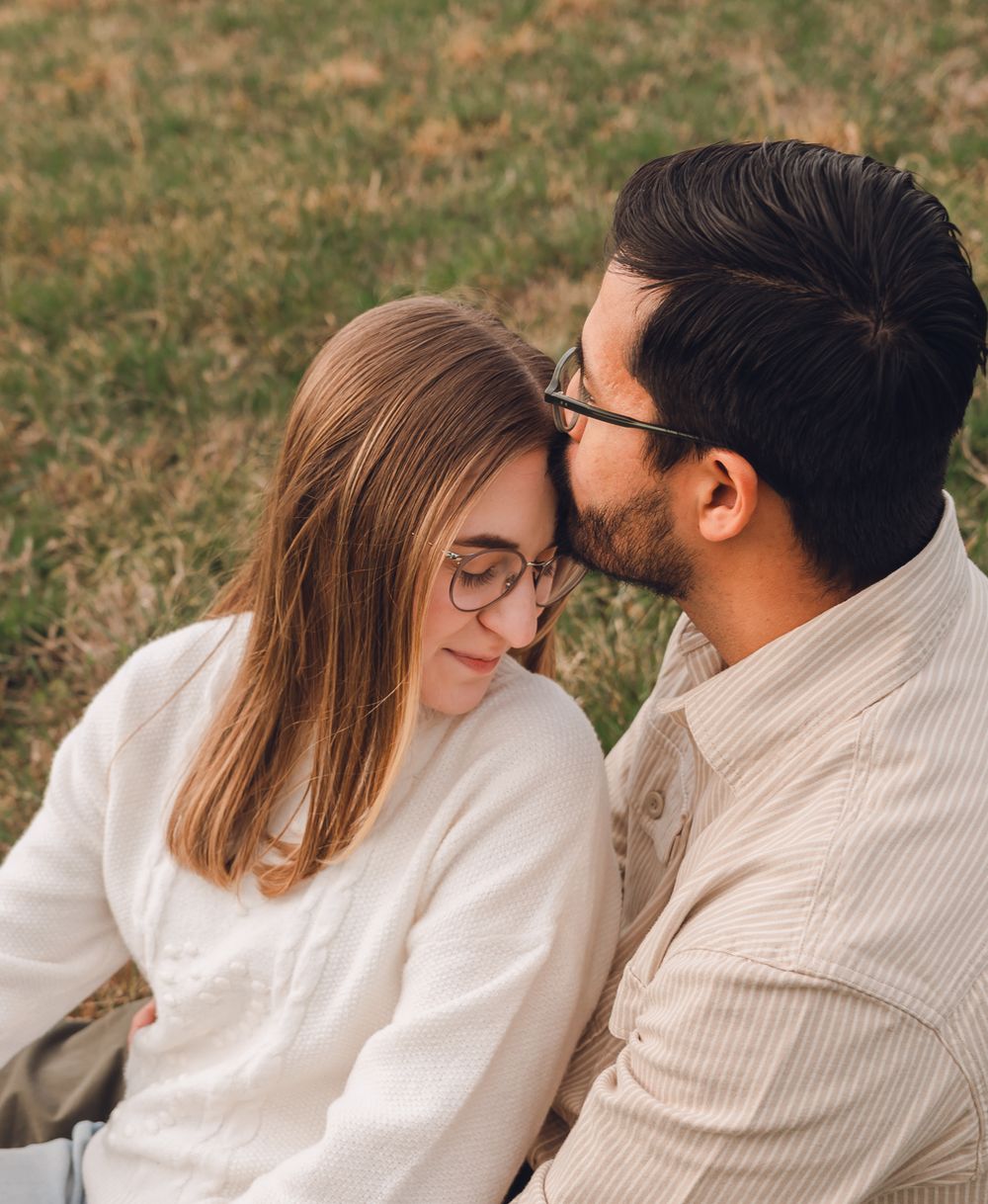 A couple shares a tender forehead kiss while sitting outdoors on green grass in casual white and beige outfits.