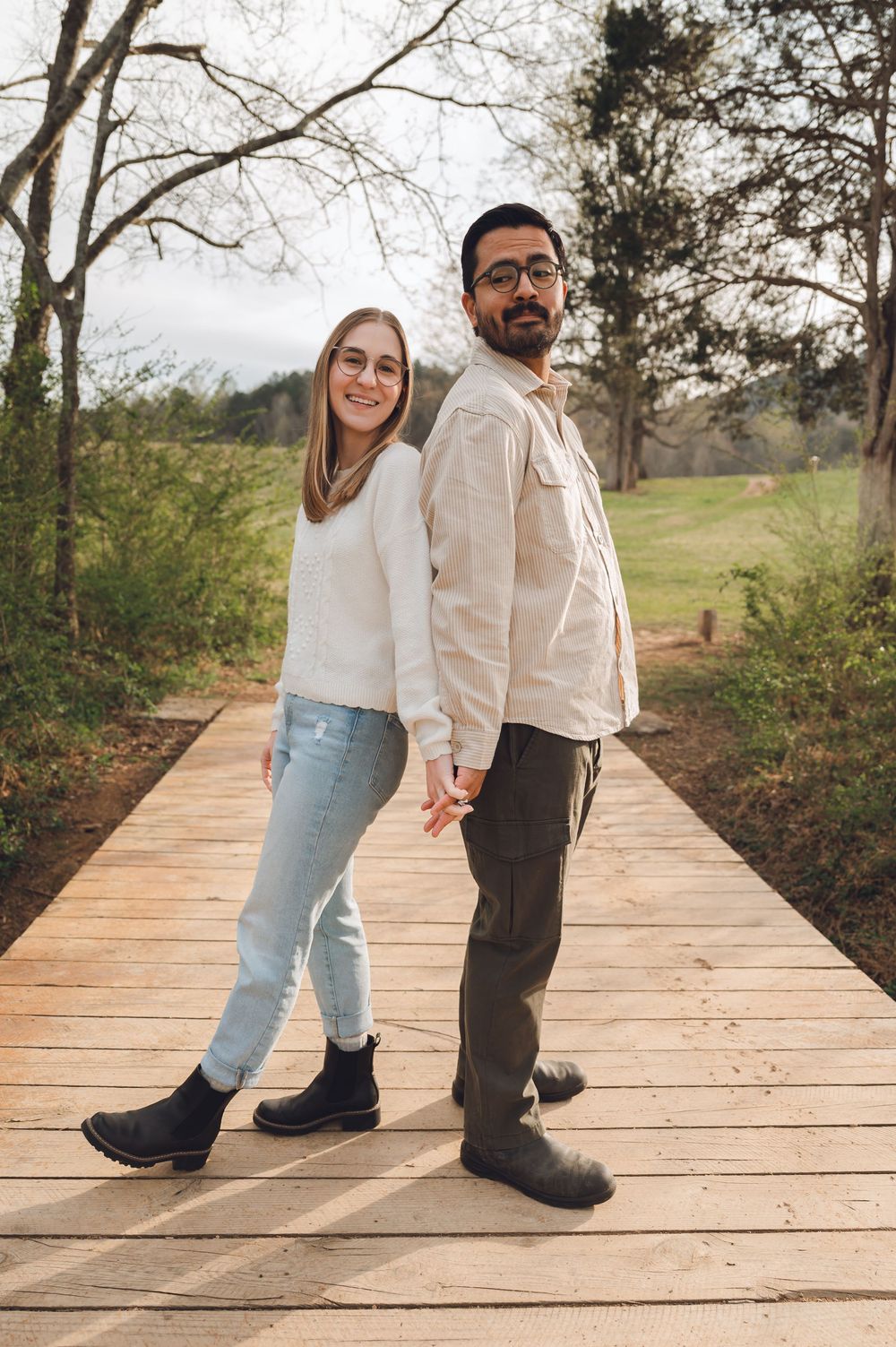 Couple holding hands and smiling on a wooden boardwalk path surrounded by trees and greenery.