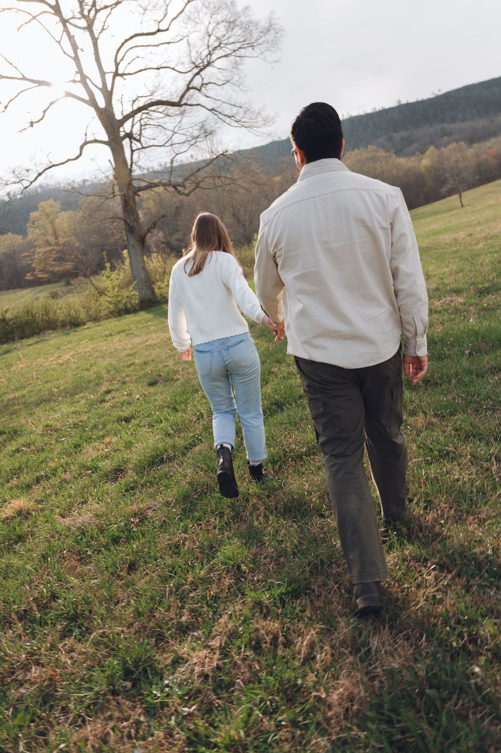 Couple holding hands walking through a lush green meadow with trees and rolling hills in the background.