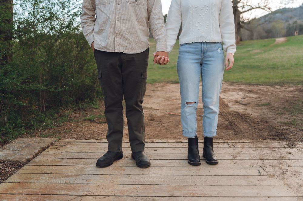 Close-up of a couple holding hands while standing on a wooden boardwalk, wearing casual outfits.
