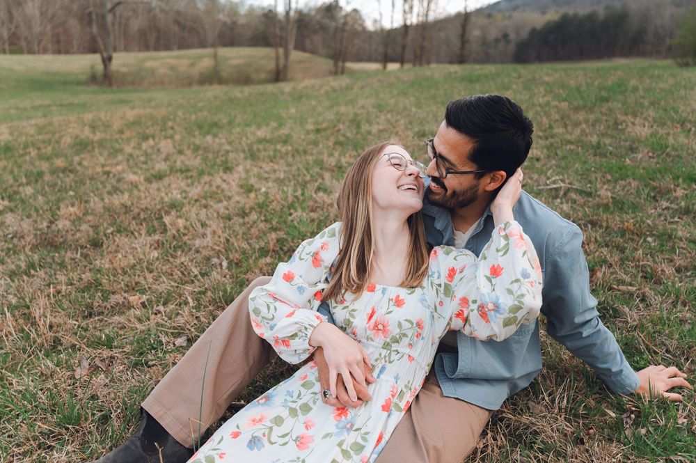 Happy couple sitting in a grassy field, woman in floral dress laughing as man leans in close on a warm day.