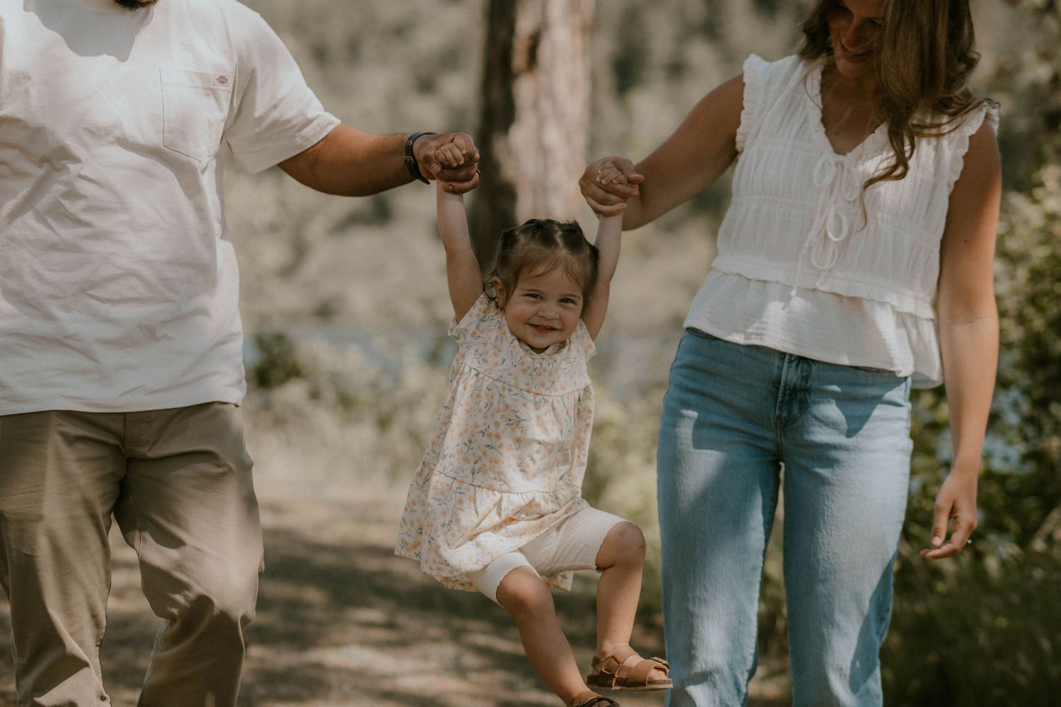 Parents swinging their happy toddler girl by her hands outdoors in a sunlit forest setting.