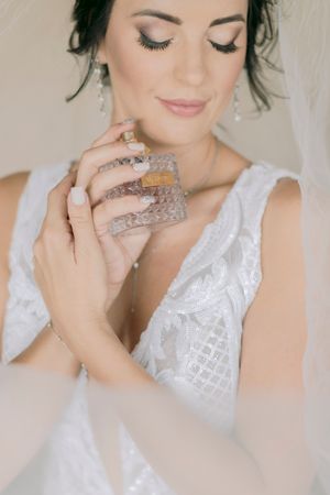 Beautiful bride in lace wedding dress applying perfume with eyes closed, soft natural light.
