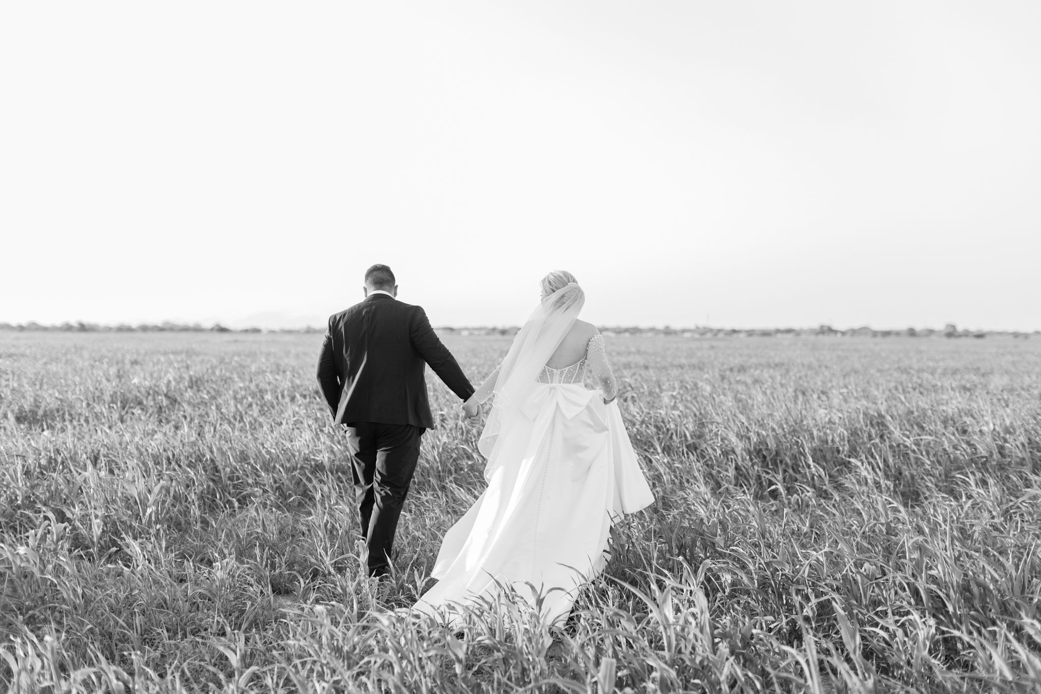 Black and white photo of bride and groom walking hand in hand through an open field.