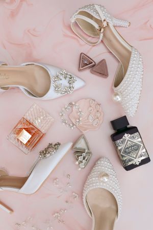 Flat lay of bridal accessories including pearl heels, white shoes, perfume, and jewelry on pink.