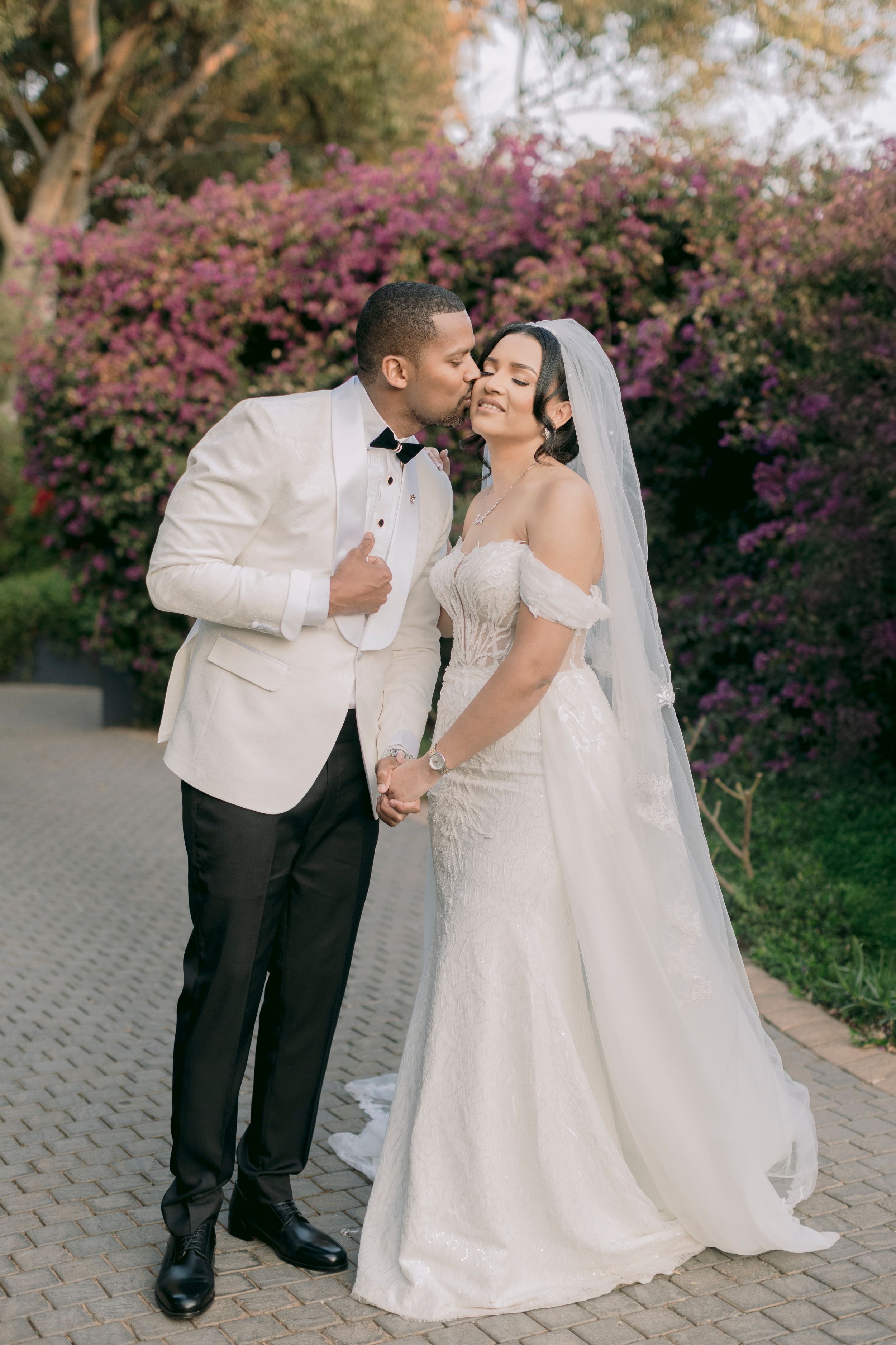 Groom in white tuxedo kissing bride in beaded off-shoulder gown outdoors with purple flowers.
