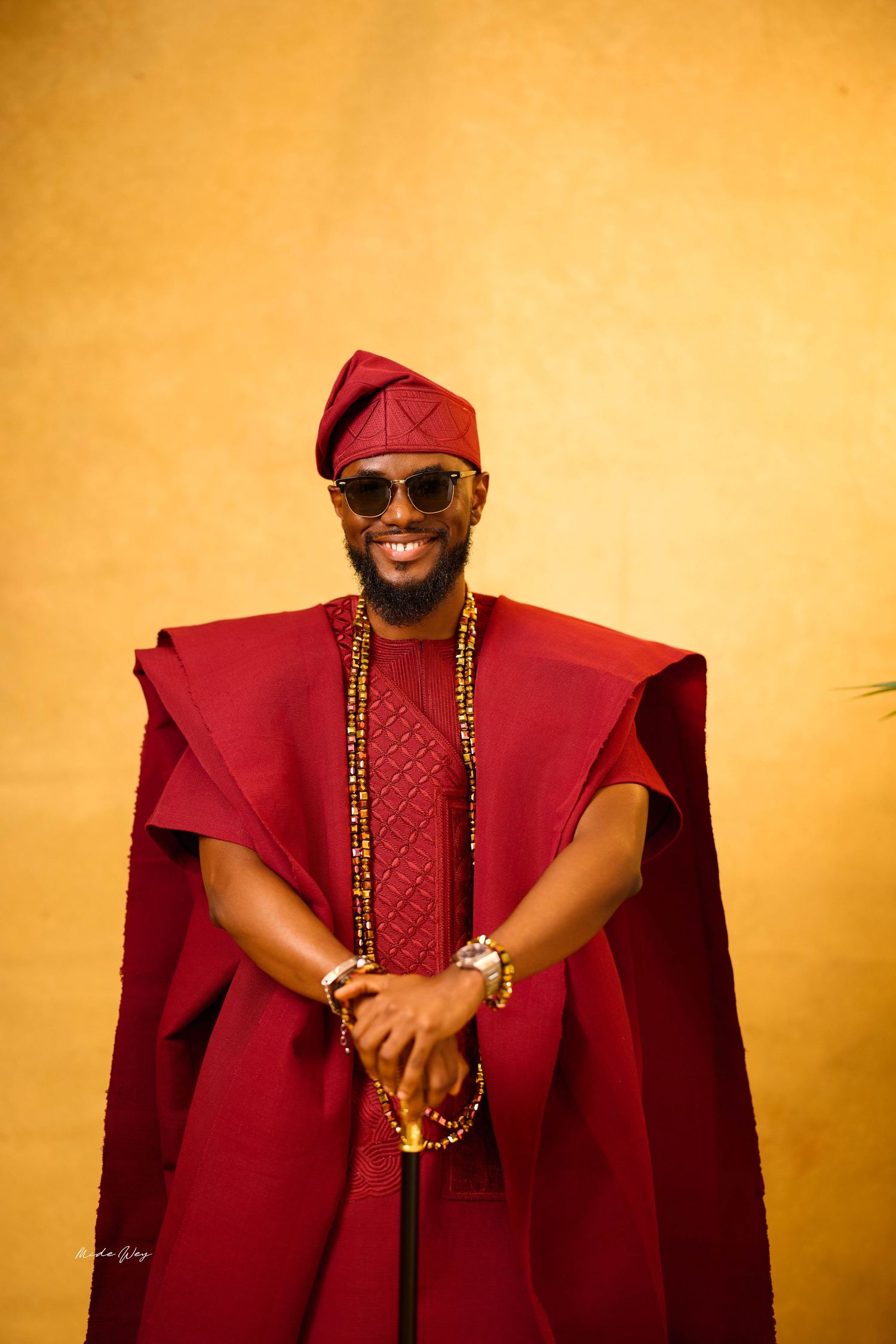 Smiling man in red Yoruba Agbada with beaded necklace and sunglasses against yellow wall.