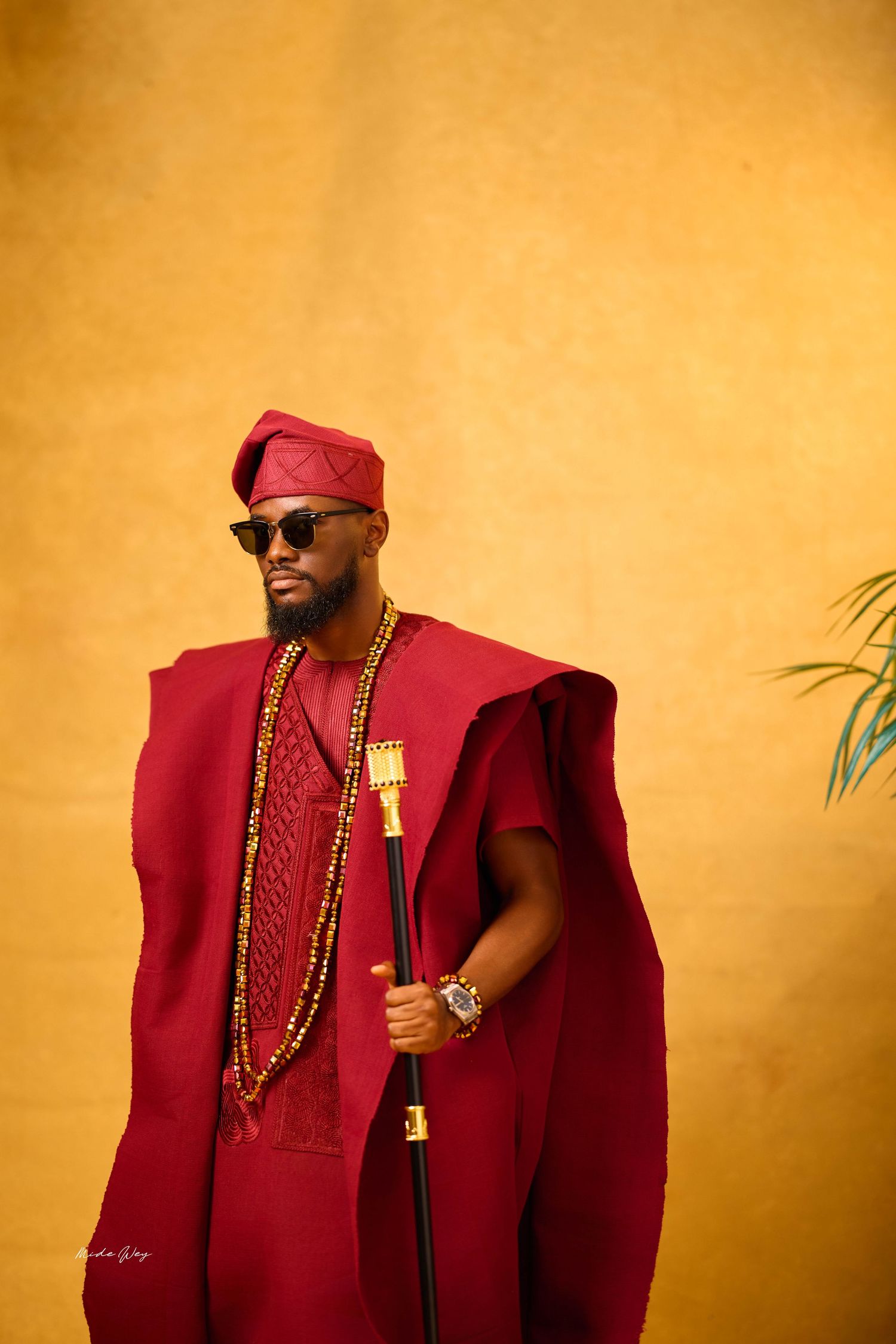 Man in red traditional Nigerian agbada outfit with matching fila cap, beaded necklaces, sunglasses, and ornate walking stick.