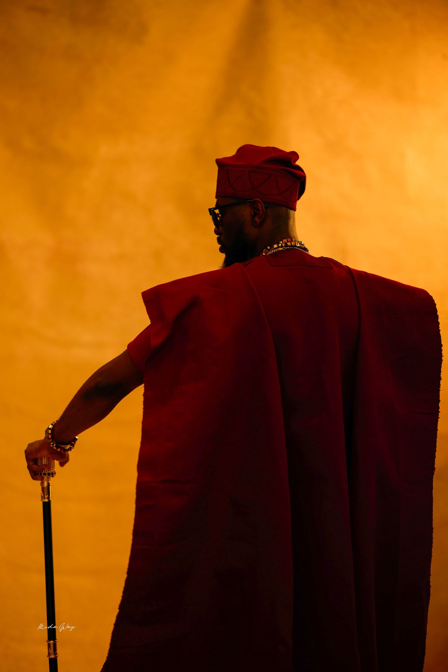 Man in red Agbada traditional Nigerian attire holding a cane, silhouetted against golden background.