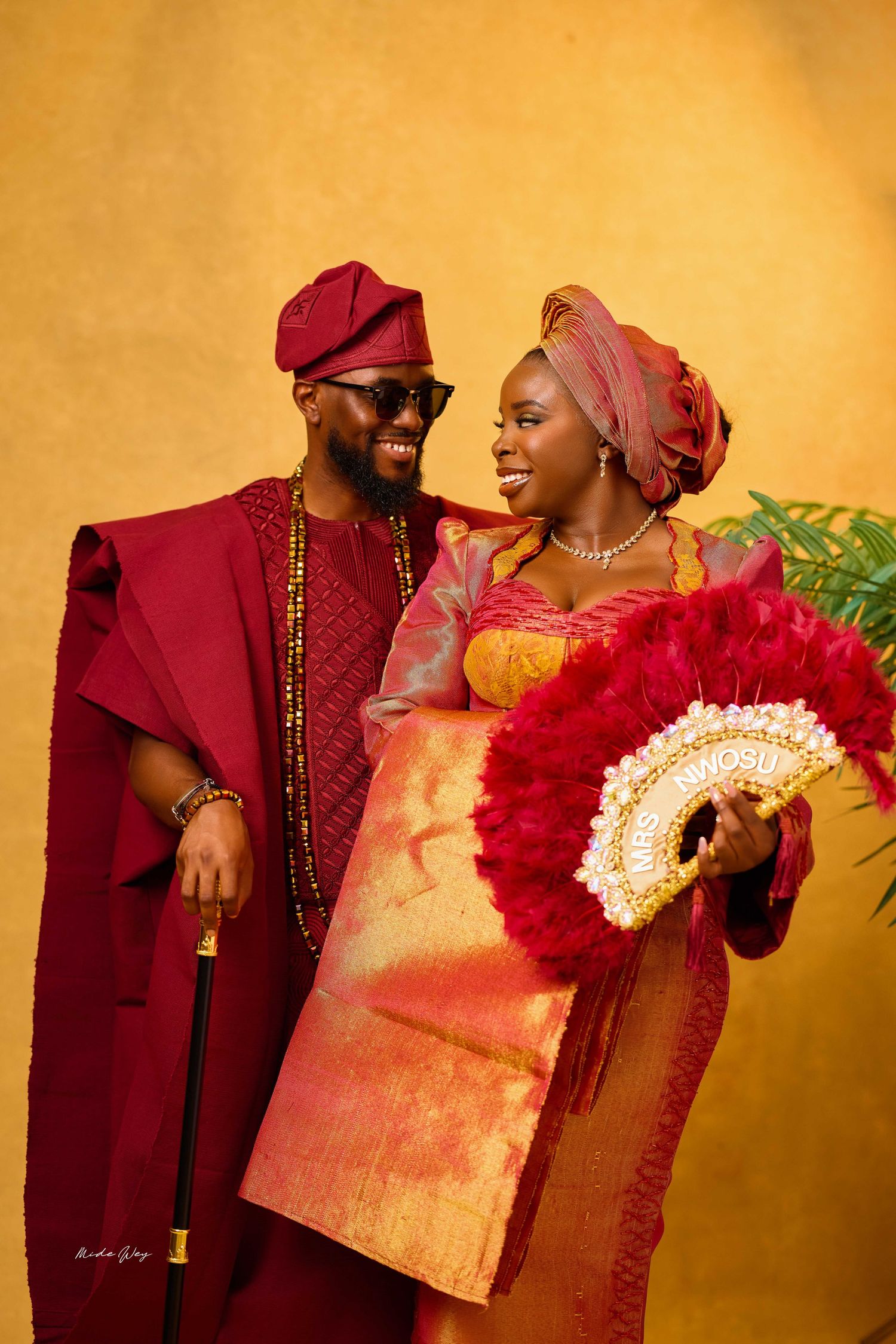 Nigerian couple in vibrant red and gold traditional Yoruba wedding attire smiling together against golden backdrop.