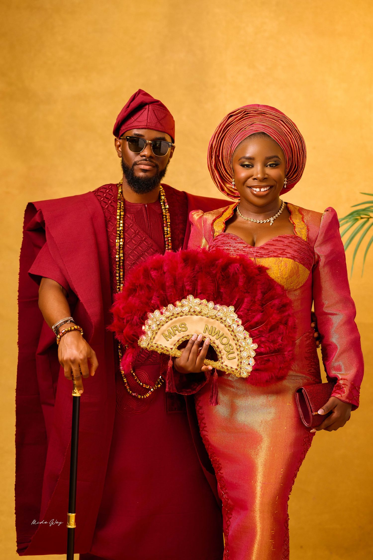 Nigerian couple in traditional Yoruba attire, man in burgundy agbada, woman in gold and red iro with gele headwrap.