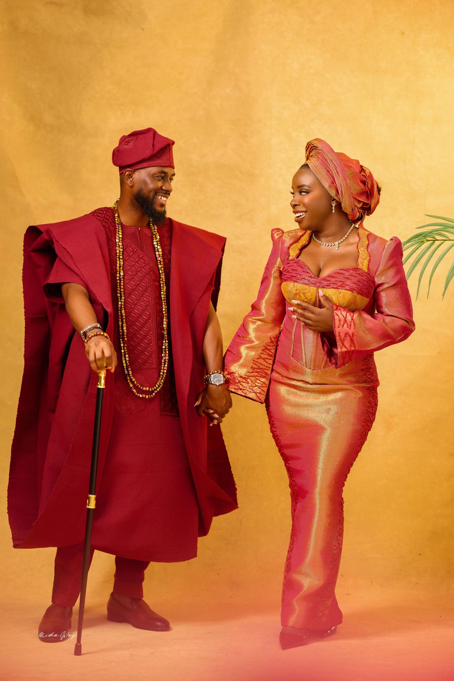 Couple in vibrant traditional Nigerian Yoruba attire, man in red agbada with cane, woman in gold and pink iro and buba.