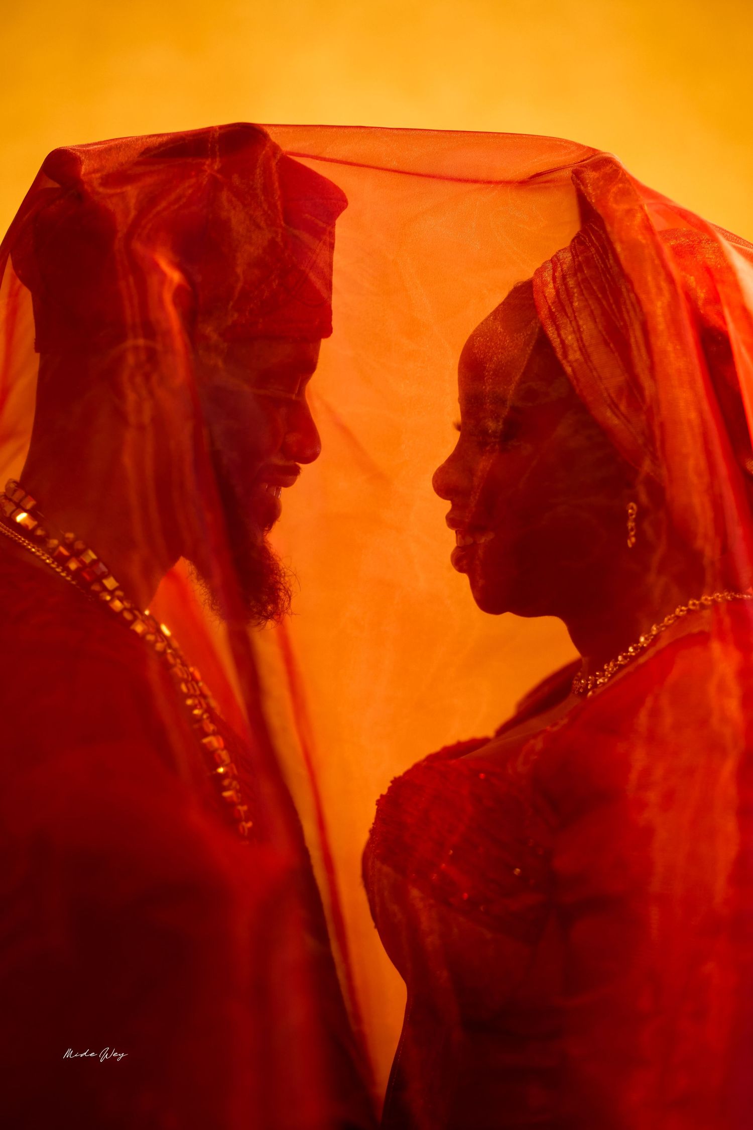 Couple in red traditional attire facing each other under a sheer red veil against a warm golden background.