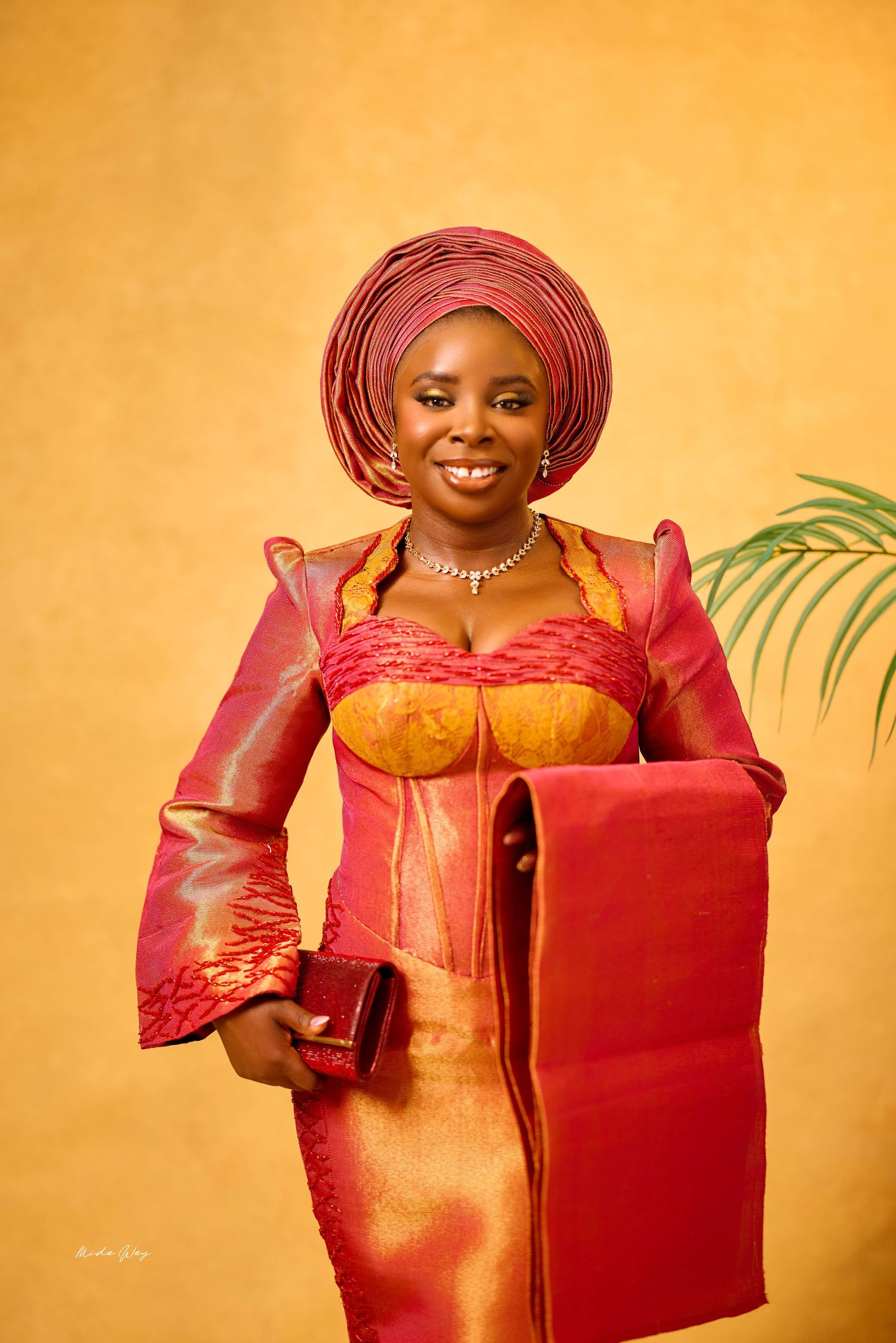 Woman in vibrant red and gold Nigerian traditional attire with gele headwrap, smiling elegantly at a portrait session.