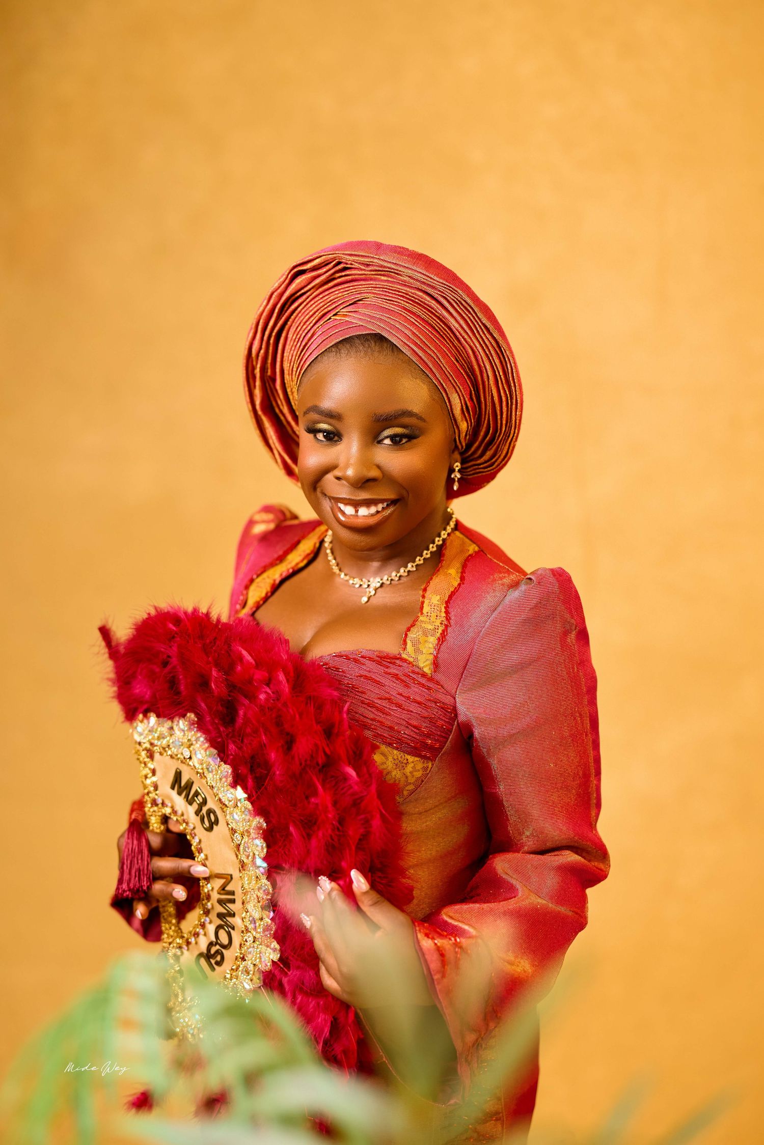 Nigerian bride in red traditional attire and gele headwrap holds decorative fan, smiling against golden background.