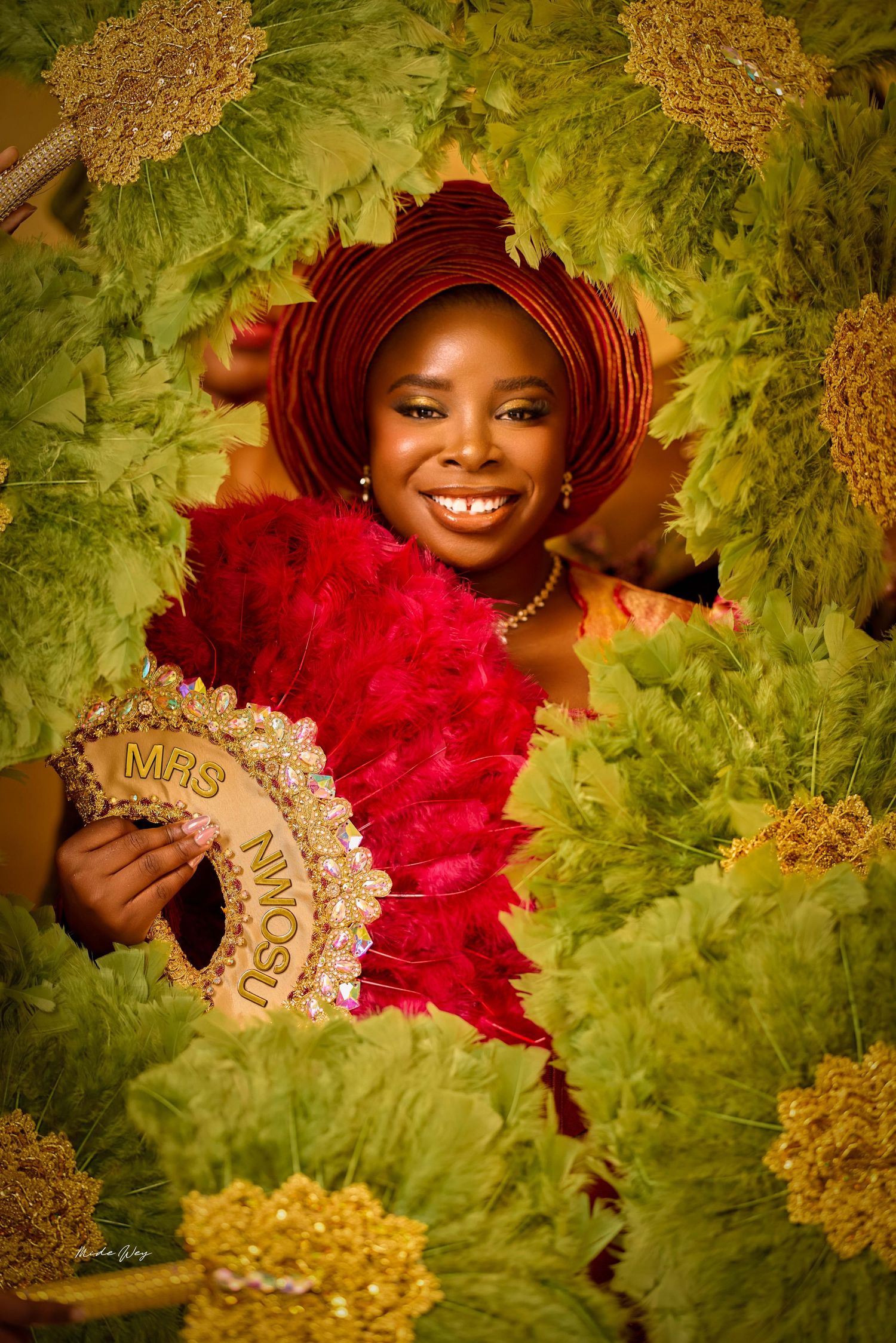 Smiling Nigerian bride in red gele headwrap holds feathered fan labeled MRS surrounded by lush green tropical foliage.