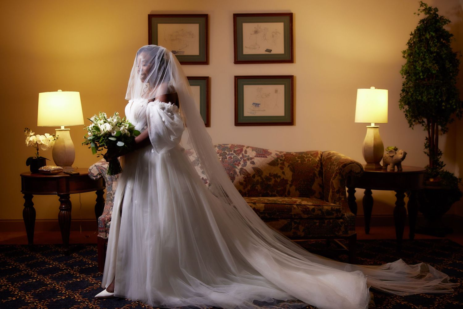 Bride in full-length white gown and veil with long train standing in warmly lit hotel suite.